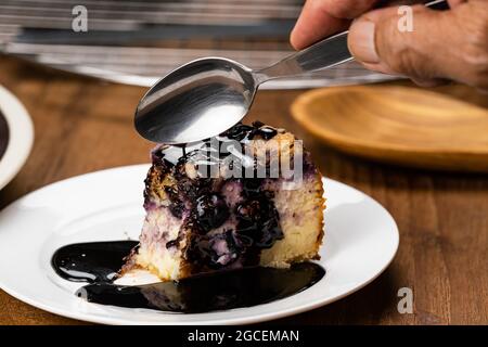 Senior Hand mit Metalllöffel essen köstliche hausgemachte Heidelbeere und bröckeln Käsekuchen Belag mit Schokolade in weißen Keramikplatte auf Holztisch Stockfoto