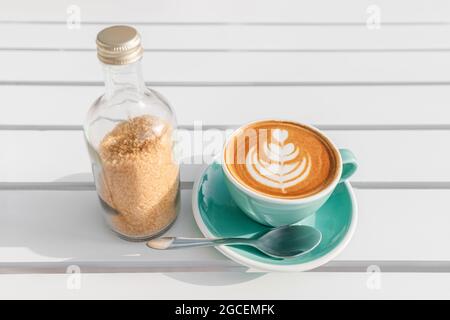 Draufsicht auf eine Tasse heißen Cappuccino mit einem einfachen Blumenmuster auf dem Schaum. Und eine Flasche Rohrzucker Stockfoto