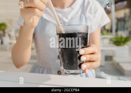 Frau trinkt ungewöhnliches kohlensäurehaltiges, funkelndes schwarzes Wasser mit Eis und Zitrone und einem Trinkhalm. Erfrischungs- und Café-Konzept Stockfoto