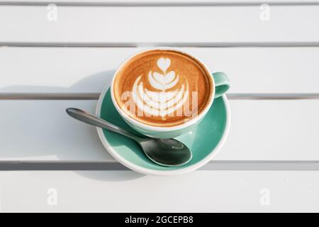 Draufsicht auf eine Tasse heißen Cappuccino mit einem einfachen Blumenmuster auf dem Schaum. Weißer Tisch in einem Sommercafé Stockfoto