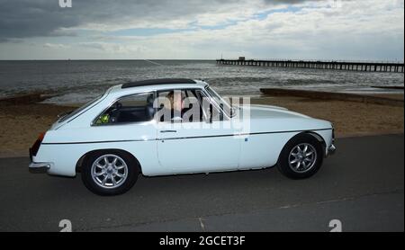 Der klassische weiße MGB GT Motor Car wird entlang der Strandpromenade mit stürmischem Himmel gefahren. Stockfoto