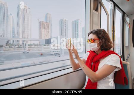 Eine Passagierin mit medizinischer Maske fährt mit öffentlichen Verkehrsmitteln und fotografiert mit ihrem Smartphone. Das Konzept der Coronavirus-Einschränkungen und qu Stockfoto