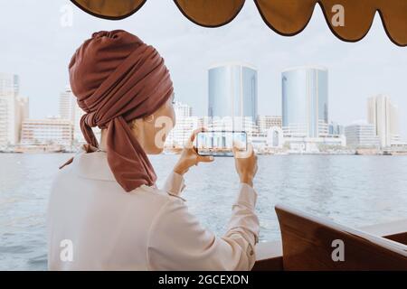 Eine glückliche asiatische Frau in einem roten Turban fotografiert auf ihrem Smartphone, während sie auf einem traditionellen Abra Dhow-Boot auf dem Dubai Creek fährt. Reisen in VAE concep Stockfoto