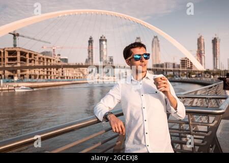 Der selbstbewusste und erfolgreiche männliche Geschäftsmann trinkt Kaffee auf dem Dubai Creek Canal Dankment mit der wunderschönen Tolerance Suspension Bridge im Hintergrund Stockfoto