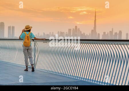 Eine glückliche Reisende Frau mit einem Hut und einem gelben Rucksack genießt einen atemberaubenden Panoramablick auf den Dubai Creek Canal und den berühmten höchsten Wolkenkratzer Bu Stockfoto