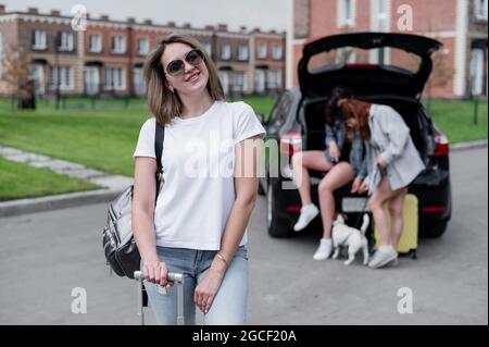 Drei Freundinnen gehen auf einen Roadtrip. Junge Frau posiert mit einem Koffer. Zwei Mädchen sitzen auf dem Kofferraum eines Autos und schauen sich eine Karte an. Stockfoto