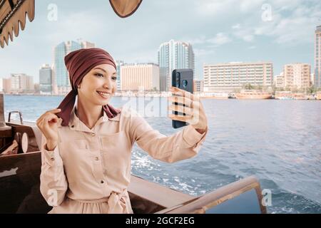 Eine glückliche asiatische Frau in einem kastanienbraunen Turban macht ein Selfie auf ihrem Smartphone, während sie auf einem traditionellen Abra Dhow-Boot auf dem Dubai Creek kreuzt. Stockfoto