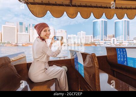 Eine glückliche asiatische Frau in einem roten Turban fotografiert auf ihrem Smartphone, während sie auf einem traditionellen Abra Dhow-Boot auf dem Dubai Creek fährt. Reisen in VAE concep Stockfoto