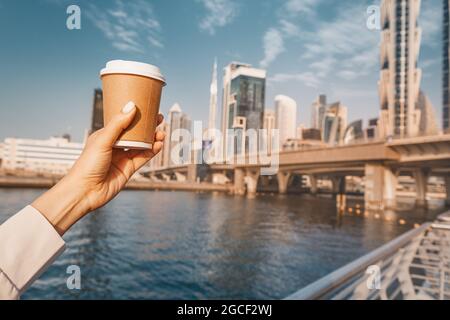Hand mit köstlichem Kaffee in Papiertasse am Dubai Canal Damm mit hohen majestätischen Wolkenkratzern im Hintergrund Stockfoto