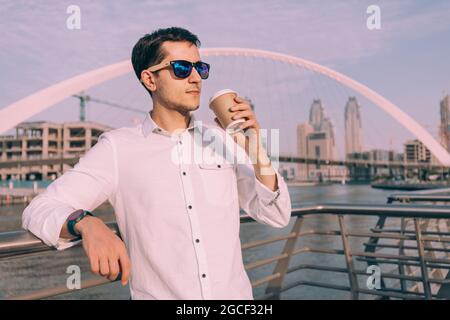 Der selbstbewusste und erfolgreiche männliche Geschäftsmann trinkt Kaffee auf dem Dubai Creek Canal Dankment mit der wunderschönen Tolerance Suspension Bridge im Hintergrund Stockfoto