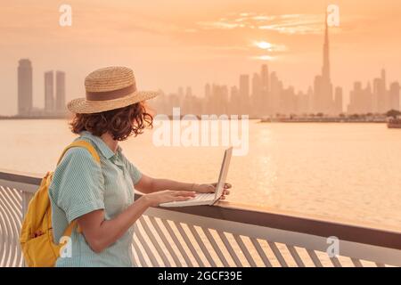 Bloggerin, Texterin oder Studentin arbeitet und studiert mit einem Laptop und bewundert den farbenfrohen Sonnenuntergang über dem Dubai Creek mit Blick auf den Wolkenkratzer Stockfoto