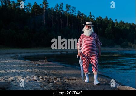 Ein älterer Mann in einem klassischen Badeanzug spaziert an einem heißen Sommertag mit einem Sonnenschirm am Strand entlang Stockfoto