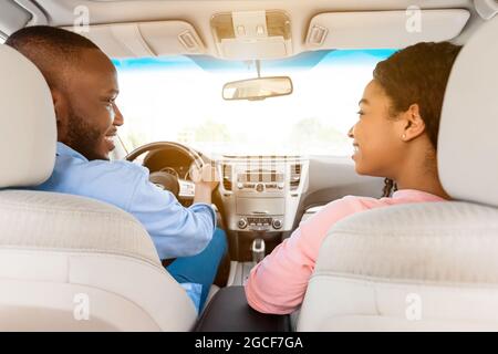 Rückansicht eines lächelnden, hübschen afroamerikanischen Mannes, der ein modernes Luxusauto fährt, einer glücklichen, schönen jungen Frau, die auf dem Beifahrersitz sitzt, lo Stockfoto