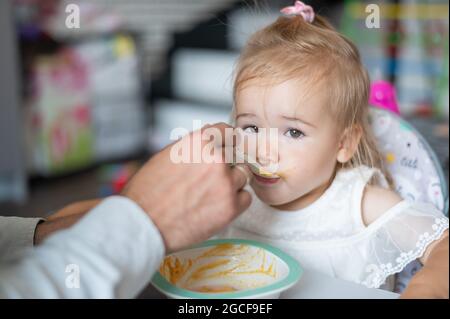 Ein fürsorglicher Mann mit Bart füttert seine kleine Tochter. Ein kleines Mädchen sitzt an einem Esstisch für Kinder und speiet mit ihrem Vater. Glückliche Vaterschaft. Stockfoto