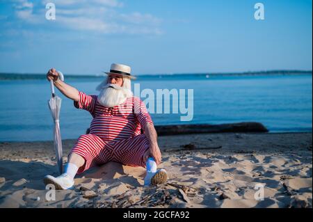 Ein alter Mann in einem klassischen Badeanzug spaziert an einem heißen Sommertag mit einem Sonnenschirm am Strand entlang Stockfoto