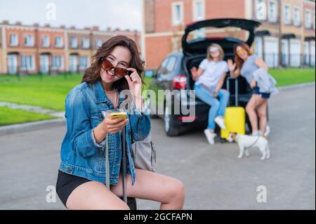 Drei Freundinnen gehen auf einen Roadtrip. Junge Frau posiert mit einem Koffer. Zwei Mädchen sitzen auf dem Kofferraum eines Autos und schauen sich eine Karte an. Stockfoto