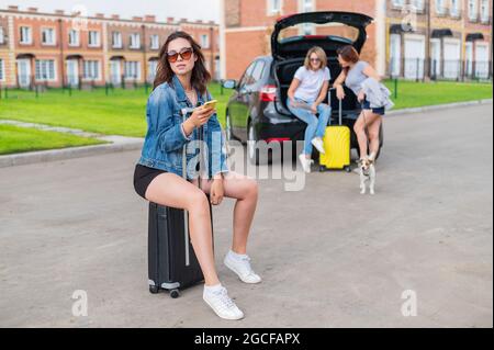 Drei Freundinnen gehen auf einen Roadtrip. Junge Frau posiert mit einem Koffer. Zwei Mädchen sitzen auf dem Kofferraum eines Autos und schauen sich eine Karte an. Stockfoto