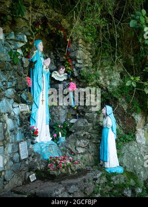 Amaga, Antioquia, Kolumbien - Juli 18 2021: Die Statuette der Jungfrau von Lourdes umgeben von Blumen und Dankesbotschaften mit einem weiteren gebrochenen Sta Stockfoto