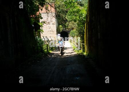 Amada, Antioquia, Kolumbien - Juli 18 2021: Lateinischer Mann und ein kleines Kind fahren an einem sonnigen Tag auf einem Motorrad durch einen dunklen Tunnel Stockfoto