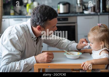 Ein fürsorglicher Mann mit Bart füttert seine kleine Tochter. Ein kleines Mädchen sitzt an einem Esstisch für Kinder und speiet mit ihrem Vater. Glückliche Vaterschaft. Stockfoto