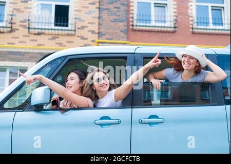 Drei fröhliche kaukasische Freundinnen schauen aus dem Autofenster. Aufgeregt Frauen gehen auf einen Roadtrip. Die Brünette Blondine und der Rotschopf gehen weiter Stockfoto