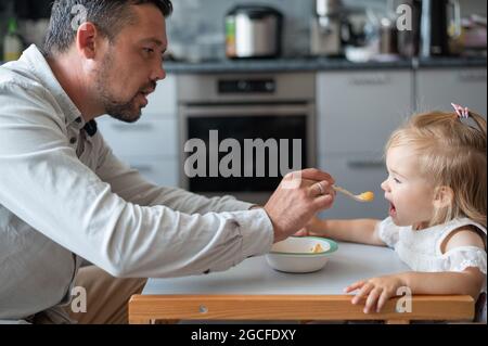 Ein fürsorglicher Mann mit Bart füttert seine kleine Tochter. Ein kleines Mädchen sitzt an einem Esstisch für Kinder und speiet mit ihrem Vater. Glückliche Vaterschaft. Stockfoto