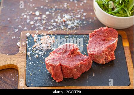 Rohes Hereford-Gras gefüttert Rinderfilet auf schwarzem slade-Brett mit Salz, Messer und einer Tasse Rucola Stockfoto