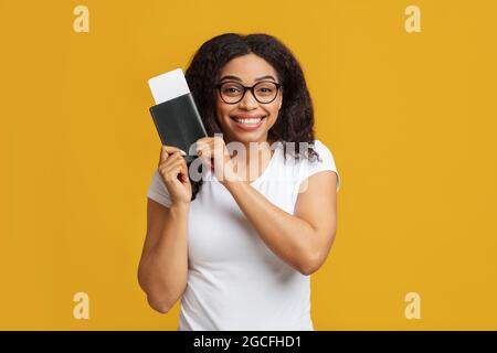 Aufgeregt afroamerikanische Frau Tourist mit Pass und Flugtickets immer bereit für den Urlaub, gelber Hintergrund. Glückliche Dame Reisende bekam ihre pla Stockfoto