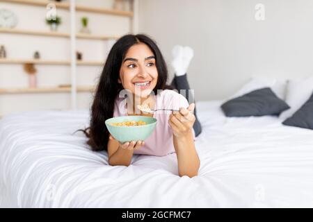 Portrait einer fittenindischen Dame, die zu Hause Müsli mit Milch auf dem Bett isst. Junge asiatische Frau genießt nahrhaftes Frühstück und hält eine ausgewogene Ernährung für das Gewicht Stockfoto