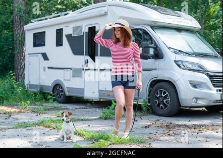 Kaukasische rothaarige Frau mit Strohhut und gestreiftem Pullover geht mit einem kleinen Hund vor ein Mobilheim. Reisen Sie mit Ihrem Haustier in einem Wohnmobil Stockfoto