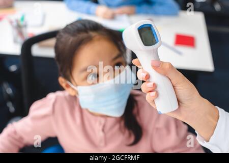 Neues Normal-, Bildungs- Und Gesundheitskonzept. Lehrer mit berührungslosem Thermometer, das die Temperatur eines kleinen asiatischen Schülers in Schutzmaske misst Stockfoto