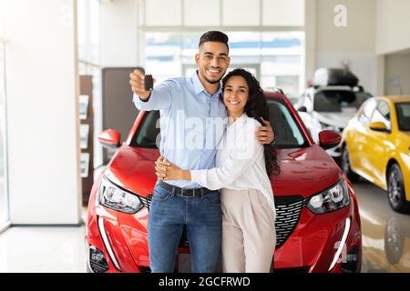 Schöne liebende muslimische Familie junger arabischer Mann und langhaarige Frau, die im Salon ein neues Auto kauft, über einem roten Luxusauto steht, umarmt und zeigt Stockfoto