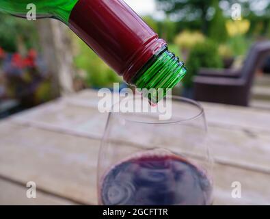 Den letzten Tropfen Rotwein aus der Flasche auf einen Glastisch aus Holz und eine verschwommene Gartenlandschaft gießen Stockfoto