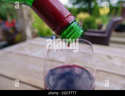 Den letzten Tropfen Rotwein aus der Flasche auf einen Glastisch aus Holz und eine verschwommene Gartenlandschaft gießen Stockfoto