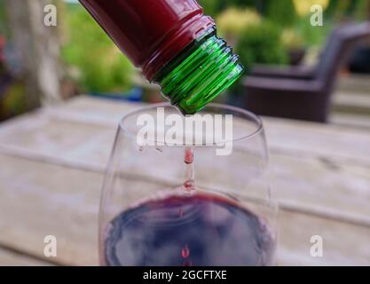 Den letzten Tropfen Rotwein aus der Flasche auf einen Glastisch aus Holz und eine verschwommene Gartenlandschaft gießen Stockfoto