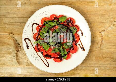 Rote Kirschtomaten und Basilikumblätter auf weißem Teller mit Balsamico-Essigsauce und Holzhintergrund schneiden Stockfoto
