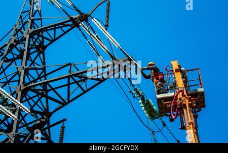 Hochspannungs-Hochspannungsleitung, Turmarbeiter mit Kran und blauem Himmel. Hydro-Linemen an Auslegerliften, die an Hochspannungstürmen arbeiten. Stockfoto