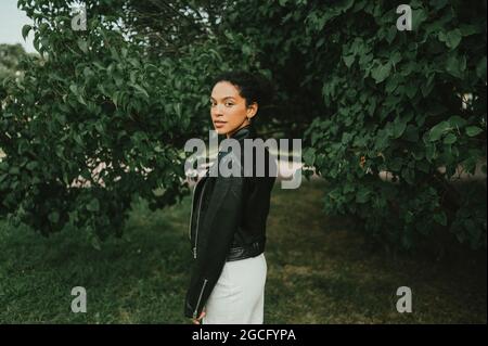 Fashion Portrait von attraktiven selbstbewussten jungen natürlichen Schönheit afroamerikanische Frau mit afro Haar in schwarzer Lederjacke posiert im Naturpark in grünem Laub. Stockfoto
