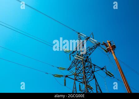 Hochspannungs-Hochspannungsleitung, Turmarbeiter mit Kran und blauem Himmel. Hydro-Linemen an Auslegerliften, die an Hochspannungstürmen arbeiten. Stockfoto