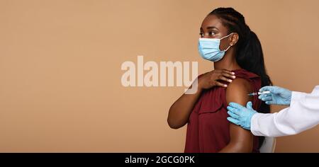 Seitenansicht einer jungen schwarzen Patientin, die eine schützende Gesichtsmaske trägt, die gegen das Coronavirus impfen kann, während eine COVID-19-Pandemie vorkam und eine Krankenschwester eine Injie machte Stockfoto