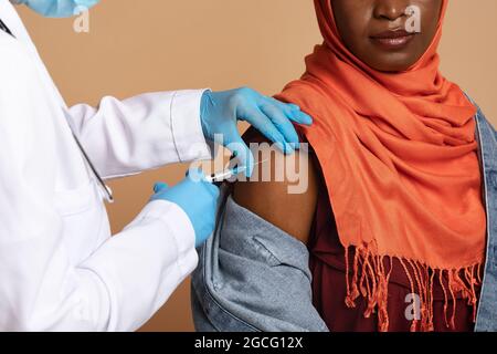 Nahaufnahme der Krankenschwesternhände in medizinischen Schutzhandschuhen, die eine intramuskuläre Injektion in die Schulter für eine muslimische schwarze Frau vornehmen, Nahaufnahme. Impfung, imm Stockfoto