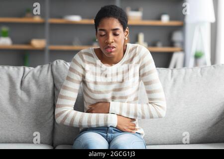 Magen-Ache. Kranke Schwarze Frau, Die Zu Hause An Akuten Bauchschmerzen Leidet, Junge Afrikanische Frau, Die Den Bauch Berührt, Während Sie Auf Der Couch Sitzt Und Menstrus Hat Stockfoto