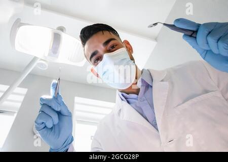 Low-Winkel-Ansicht der jungen arabischen Zahnarzt hält zahnärztliche Werkzeuge, bereit für die Untersuchung, professionelle Nahost Stomatologe Arzt trägt medizinische Maske c Stockfoto