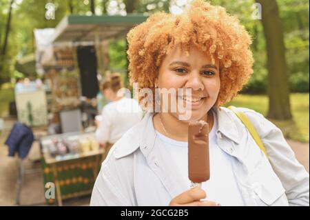 Lifestyle-Porträt von lächelnden jungen natürlich schönen plus size afroamerikanischen Frau mit afro roten Haaren essen Eis Schokolade Popsicle im öffentlichen Park. Vielfalt und körperpositives Konzept Stockfoto