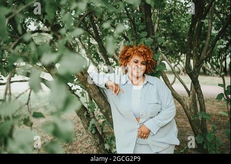 Fashion Portrait von jungen natürlich schönen plus size afroamerikanische Frau in hellen Hosenanzug mit afroroten Haaren posiert in Natur Parkland in grünem Laub. Vielfalt und körperpositives Konzept Stockfoto