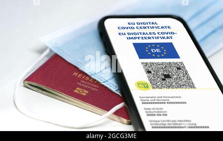 Digitales EU-COVID-Zertifikat mit dem QR-Code auf dem Bildschirm eines Mobiltelefons über einer OP-Maske und einem deutschen Reisepass. Immunität gegen Covid-1 Stockfoto