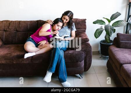 Mexikanisches Teenager-Mädchen mit Down-Syndrom und ihre Mutter mit einem Buch zu Hause, im Behindertenkonzept in Lateinamerika Stockfoto