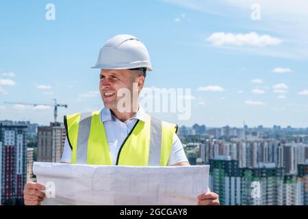 Ein Mann in einem weißen Helm hält den Bauplan fest und inspiziert die Baustelle Stockfoto