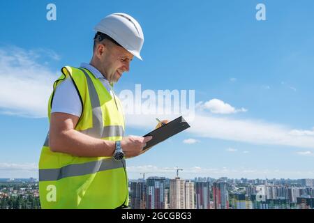 Lächelnder Mann in weißem Helm auf der Baustelle schreibt Notizen auf der Zwischenablage Stockfoto