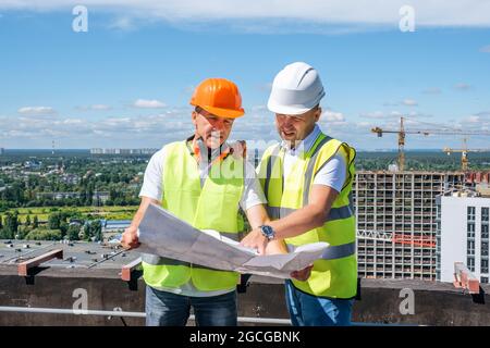 Männer in einem weißen Helm halten einen Bauplan und inspizieren die Baustelle Stockfoto